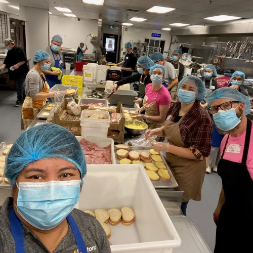 Group photo of multiple volunteers working in a kitchen serving sandwhiches and pausing in thier work to take a pictures.